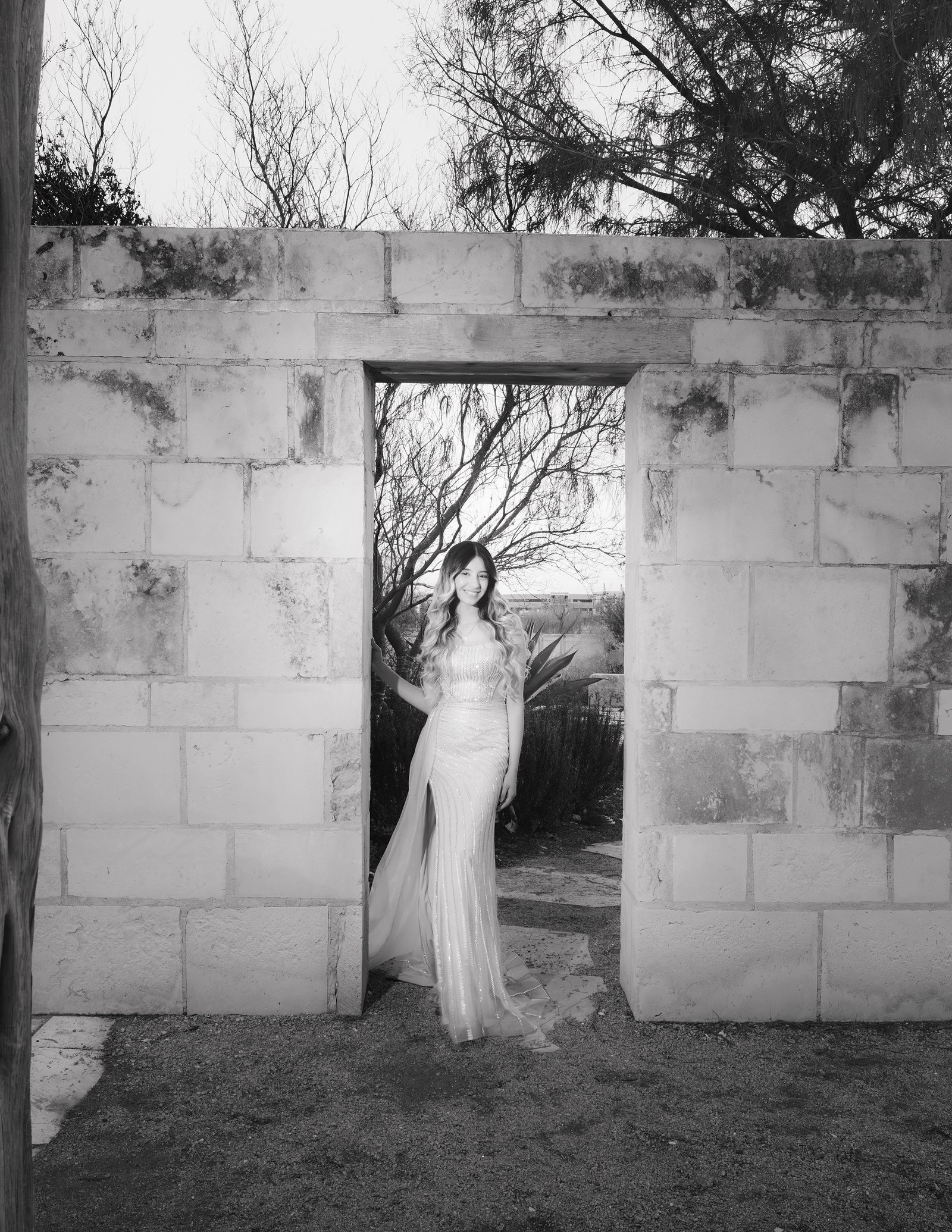 Teresa Vitela senior Black and white portrait - elegant young woman in long beige dress posing against cantera stone wall Texas A&M Gardens College Station Texas.