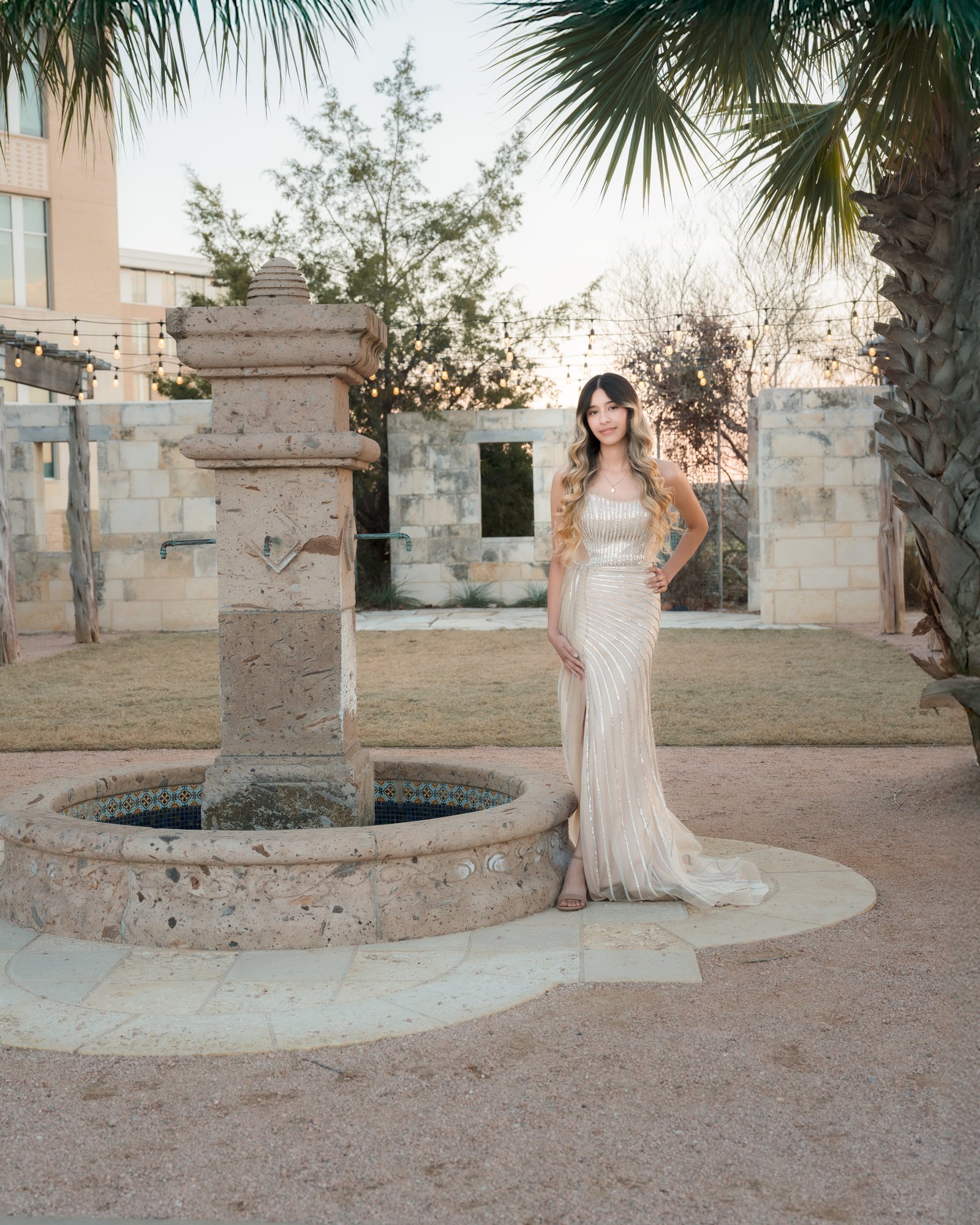 Teresa Vitela senior portrait - elegant young woman in long beige dress posing near fountain Texas A&M Gardens College Station Texas