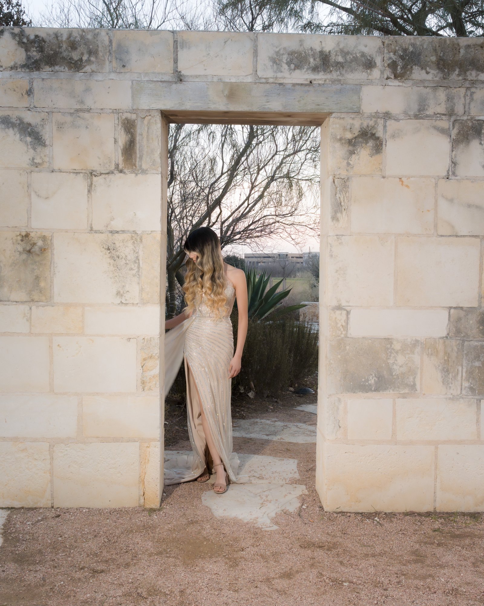 Teresa Vitela luxury senior session - magazine worthy portrait young woman in beige gown emerging through cantera stone door Texas A&M Gardens College Station Texas