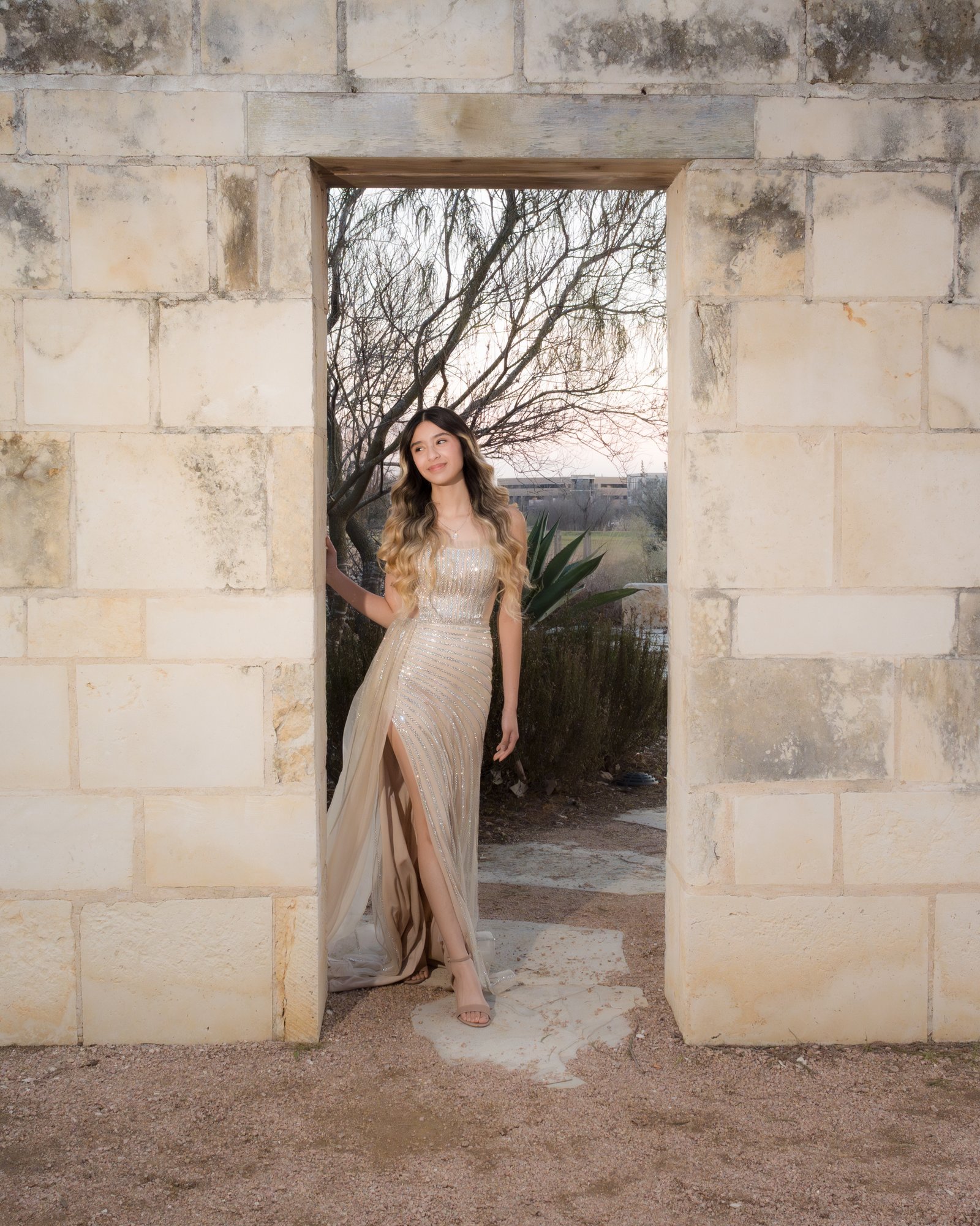 Teresa Vitela senior portrait - elegant young woman in long beige dress walking through ornate cantera stone doorway Texas A&M Gardens College Station Texas