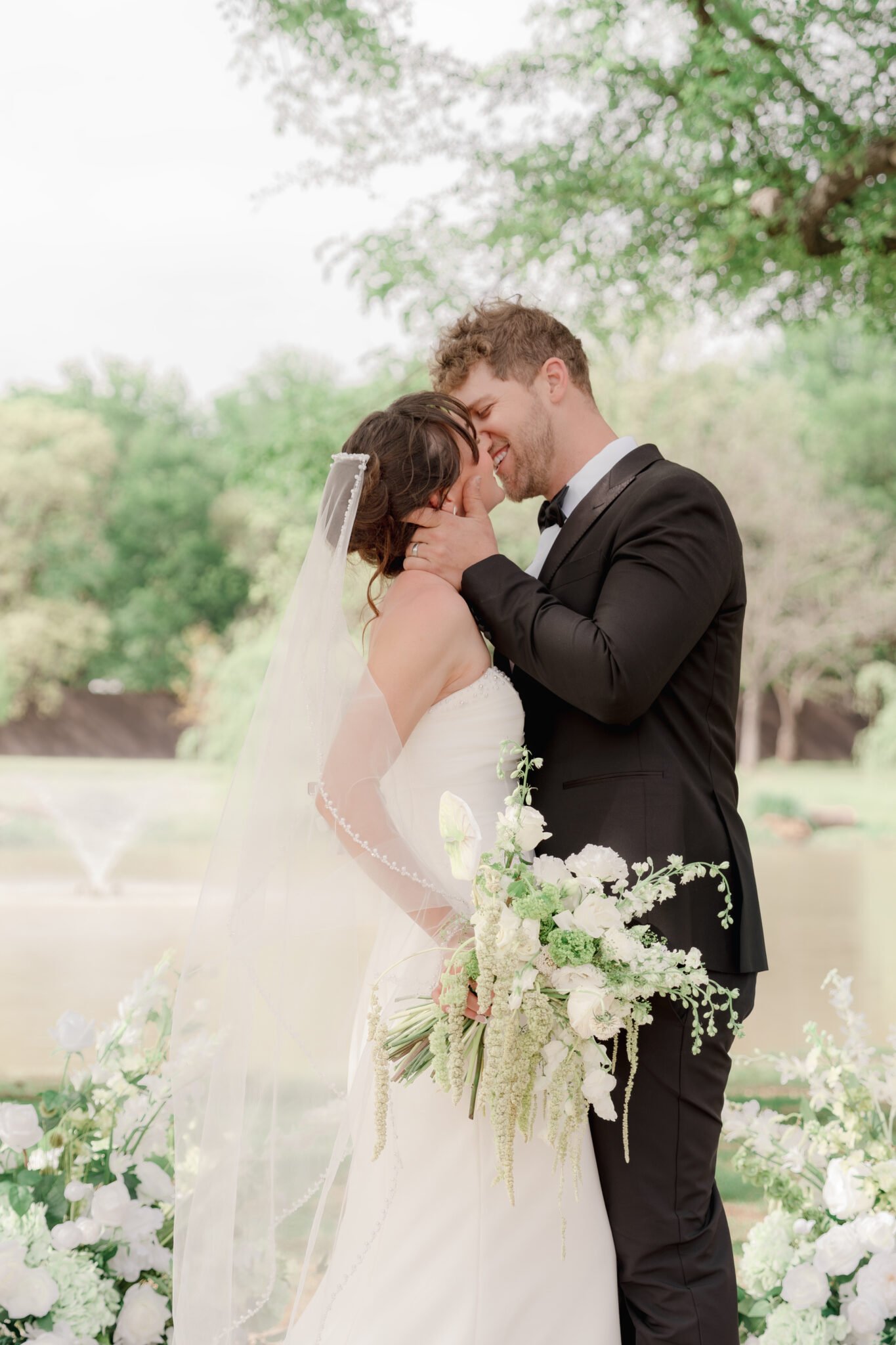 Bride and groom sharing their first kiss at the altar, surrounded by elegant white floral pillars and a scenic outdoor background.