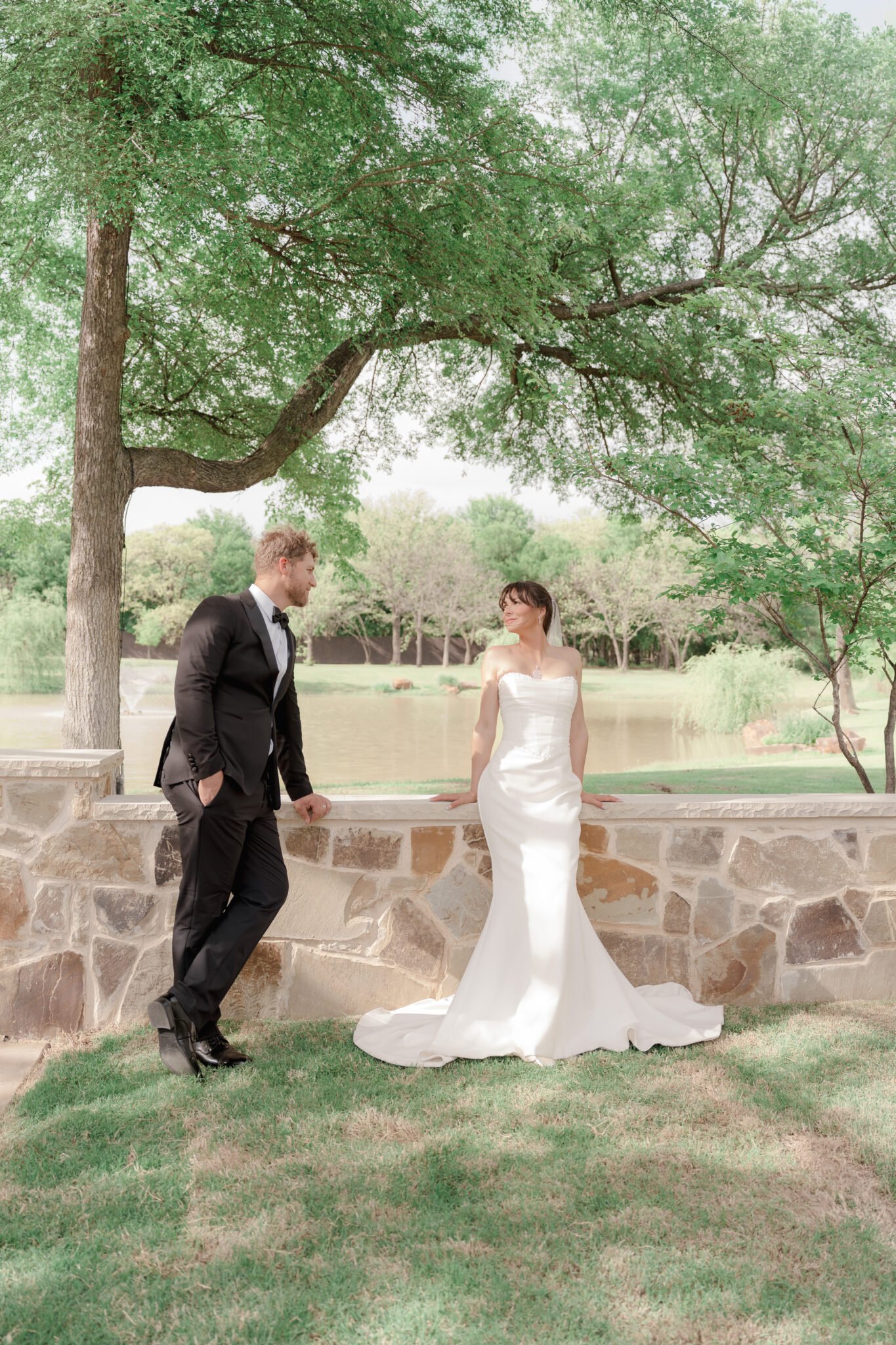 Bride and groom walking hand-in-hand past a classic stone wall in a luxury estate garden.