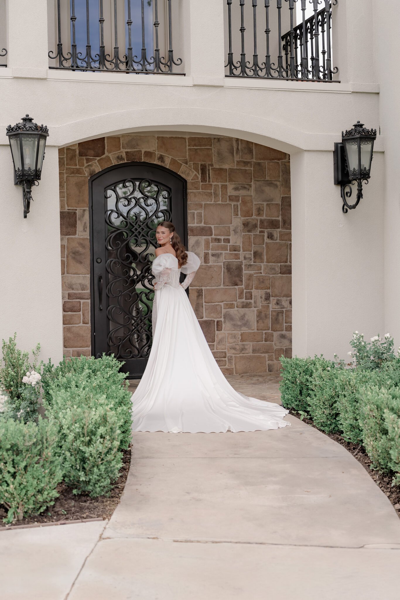A bride in a couture lace gown and long veil posing in a historic stone archway entrance.