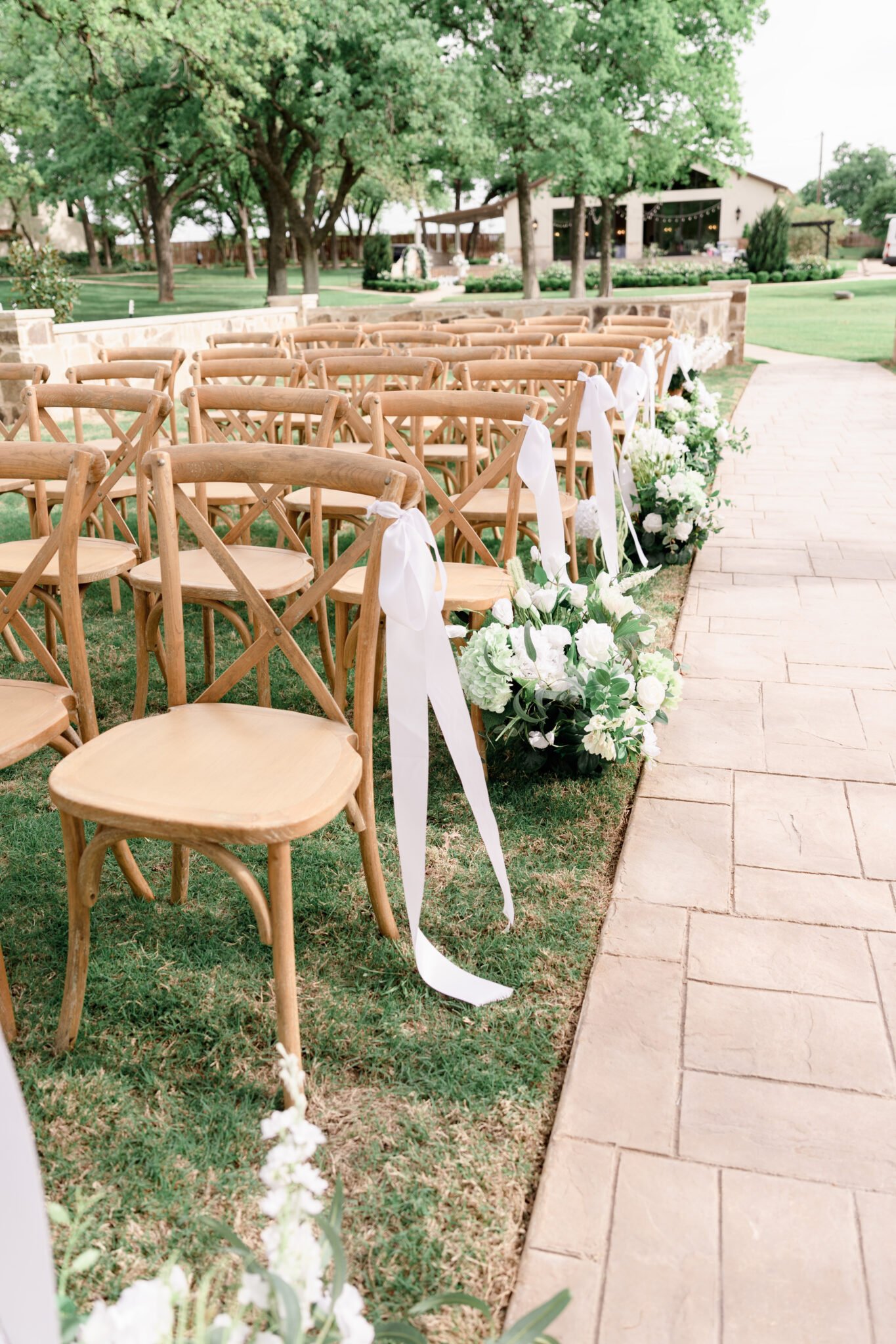 Outdoor wedding ceremony setup with wooden cross-back chairs and elegant white floral arrangements.