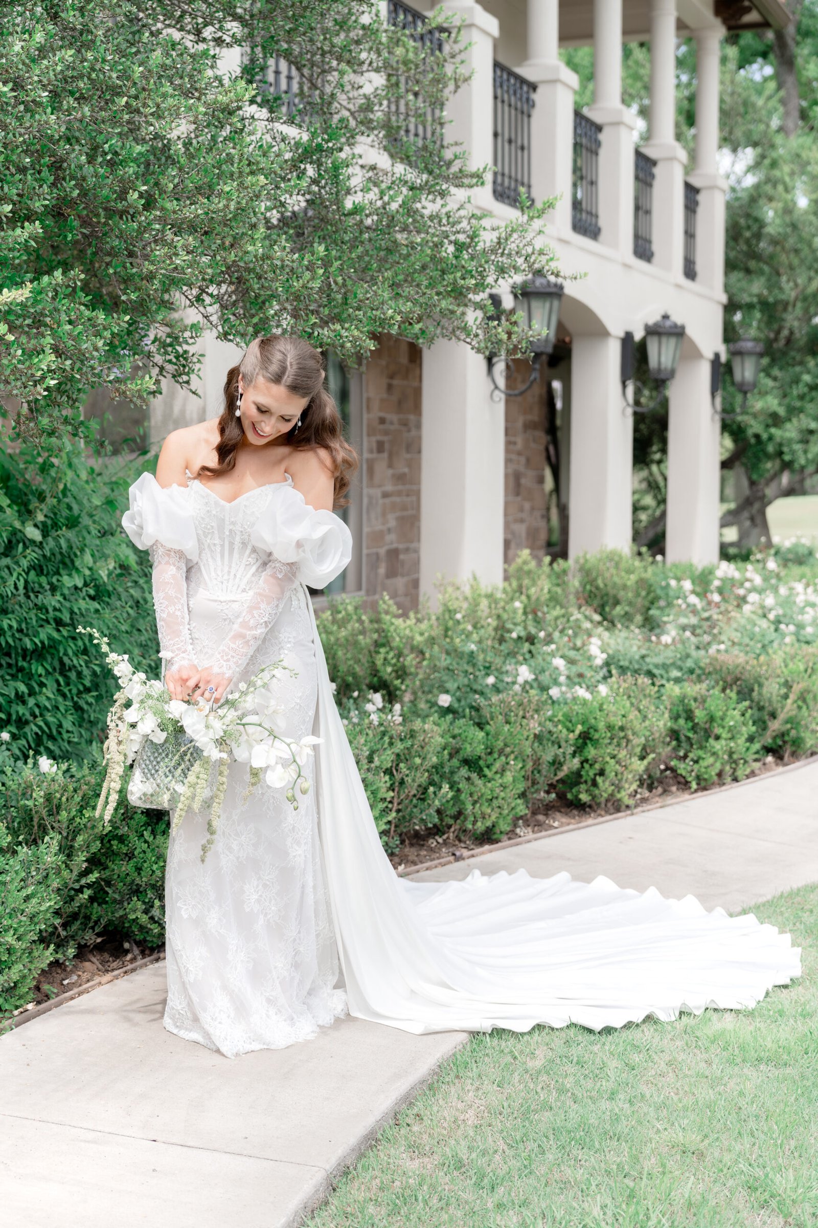 Fine art bride in off-shoulder lace gown walking along stone path in front of Tuscan villa venue in Dallas Texas
